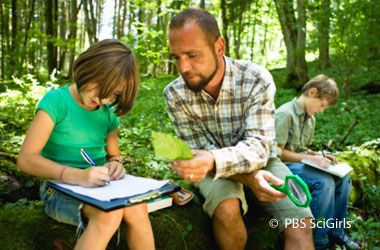 two children writing on clipboards and man showing one of them a leaf, they are in the woods