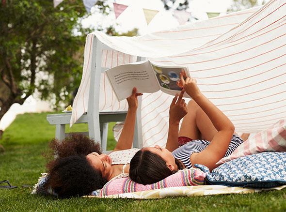 a mother and daughter lying on the ground reading a book with a sheet tented over them