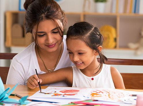 young woman sits next to young girl who is painting at a table