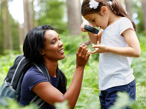 a woman holds up a pinecone which a young girl examines with a magnifying glass